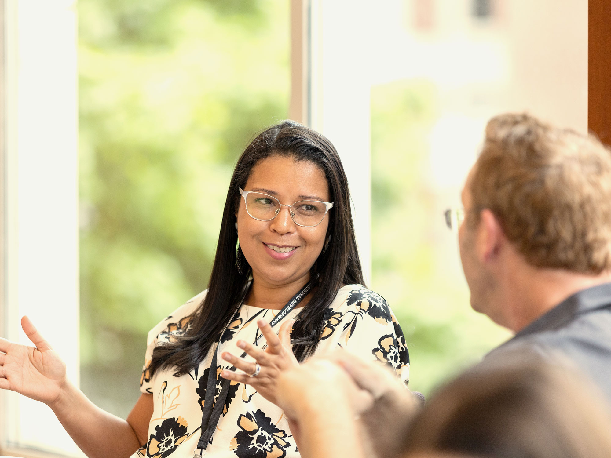 A woman with dark hair and glasses speaking cheerfully to a man. Both are sitting by brightly lit windows with trees out of focus.