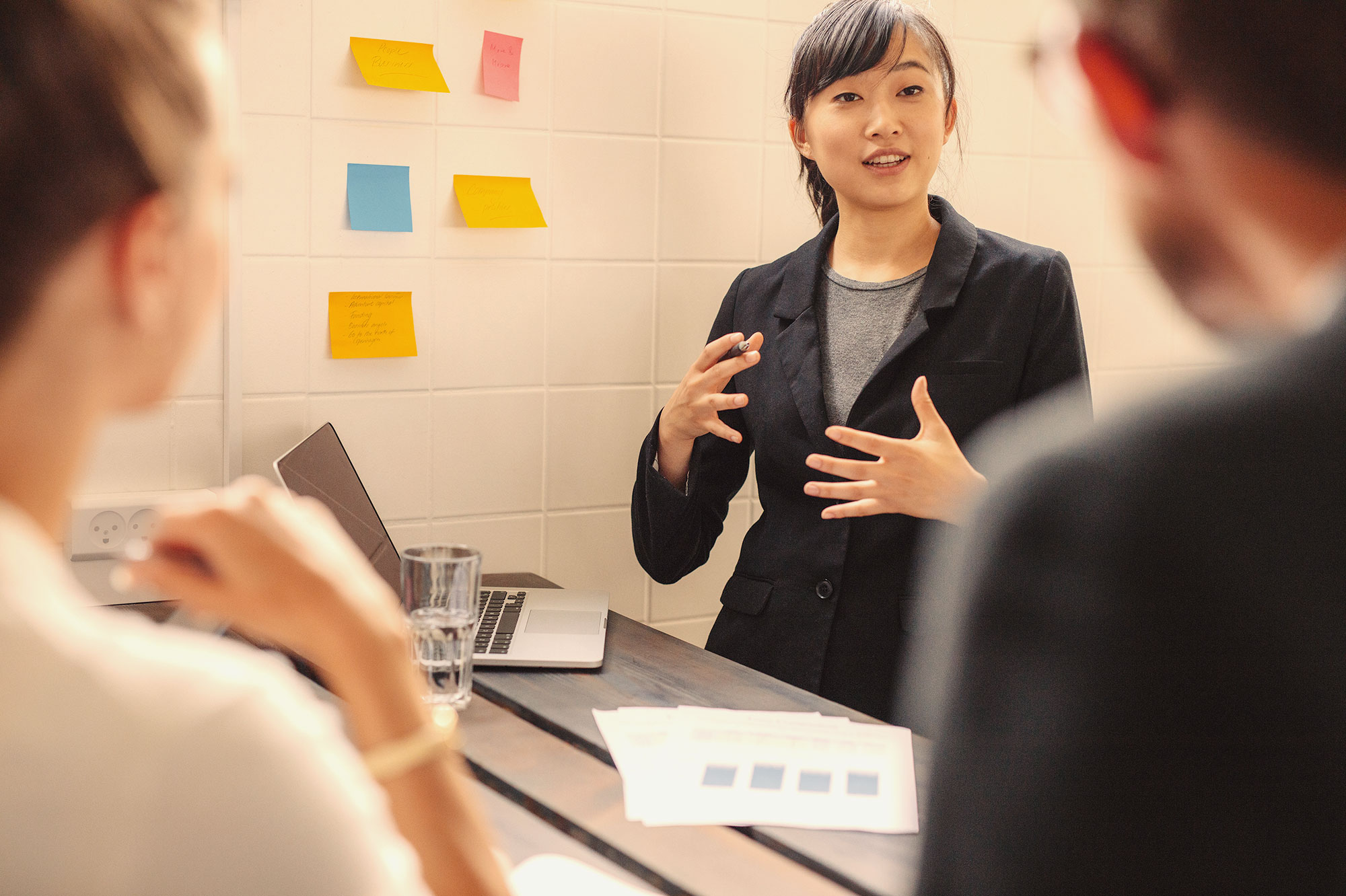 A woman stands in front of a white board with colored sticky notes explaining something (successful change initiatives)to two colleagues