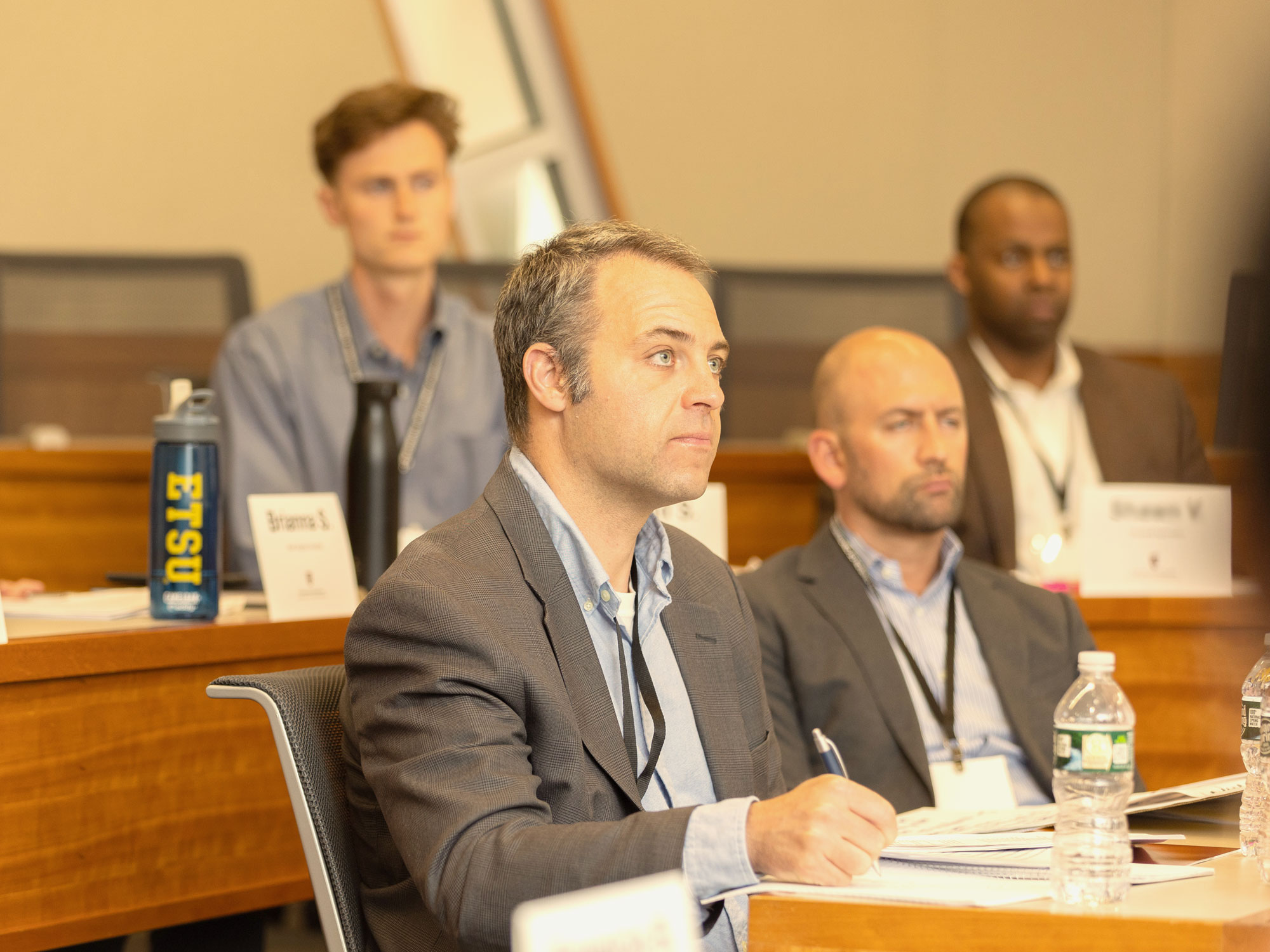 A man in a gray suit writing notes on a piece of paper. While a group of 3 men in professional attire listen to the presentation intently