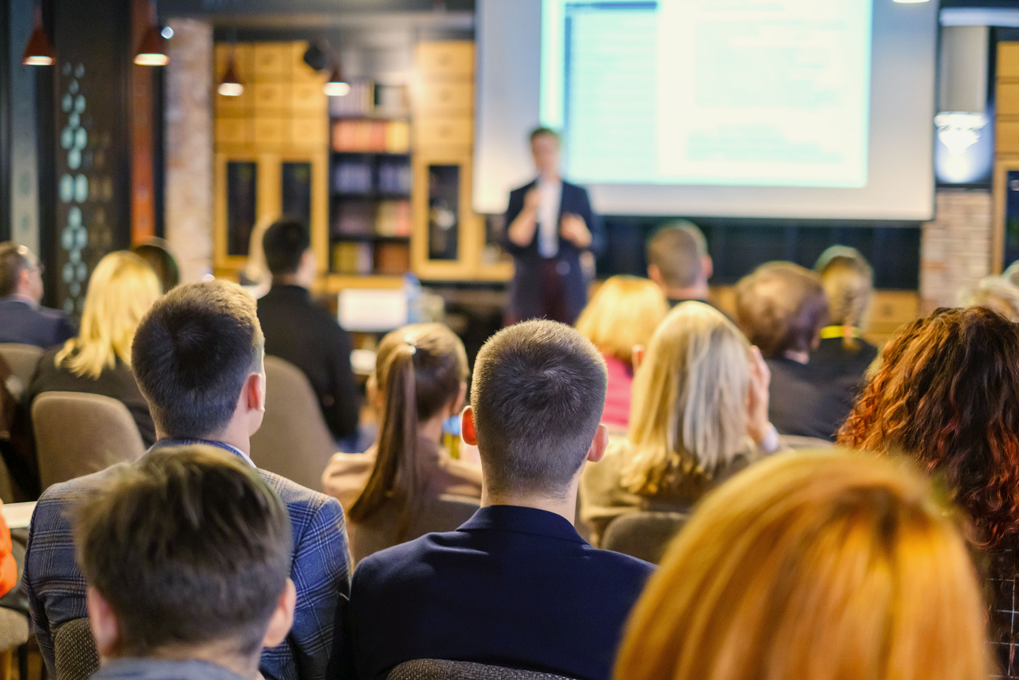 An instructor stands in front of a screen in front of a classroom.