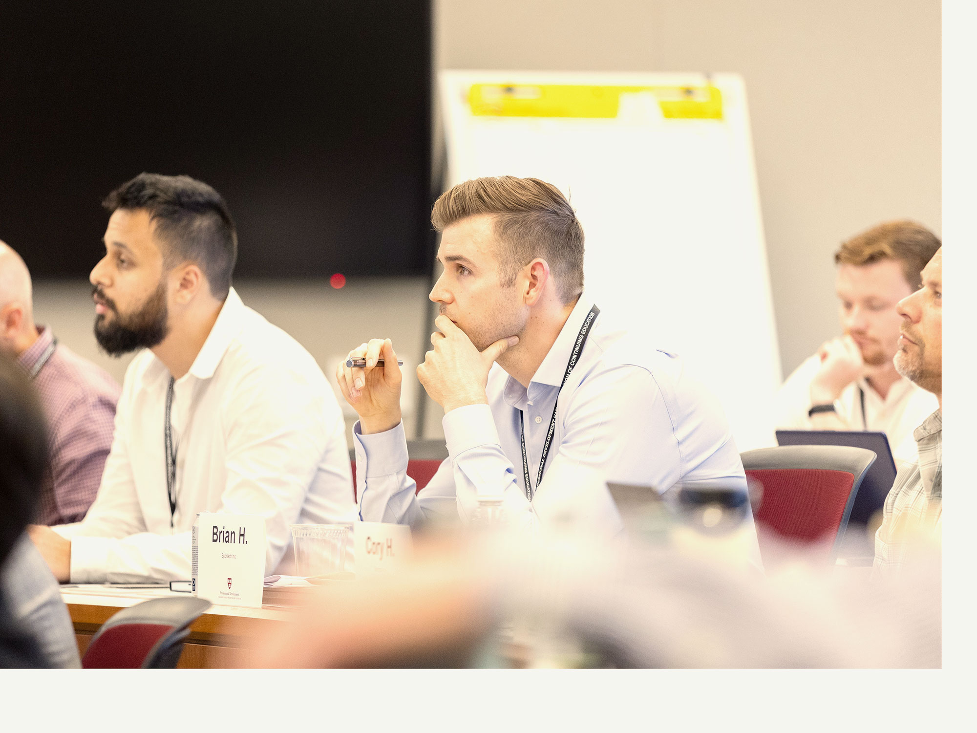 A group of adult learners sit in a classroom listening attentively. The main subject has their hand over their mouth, in deep thought.