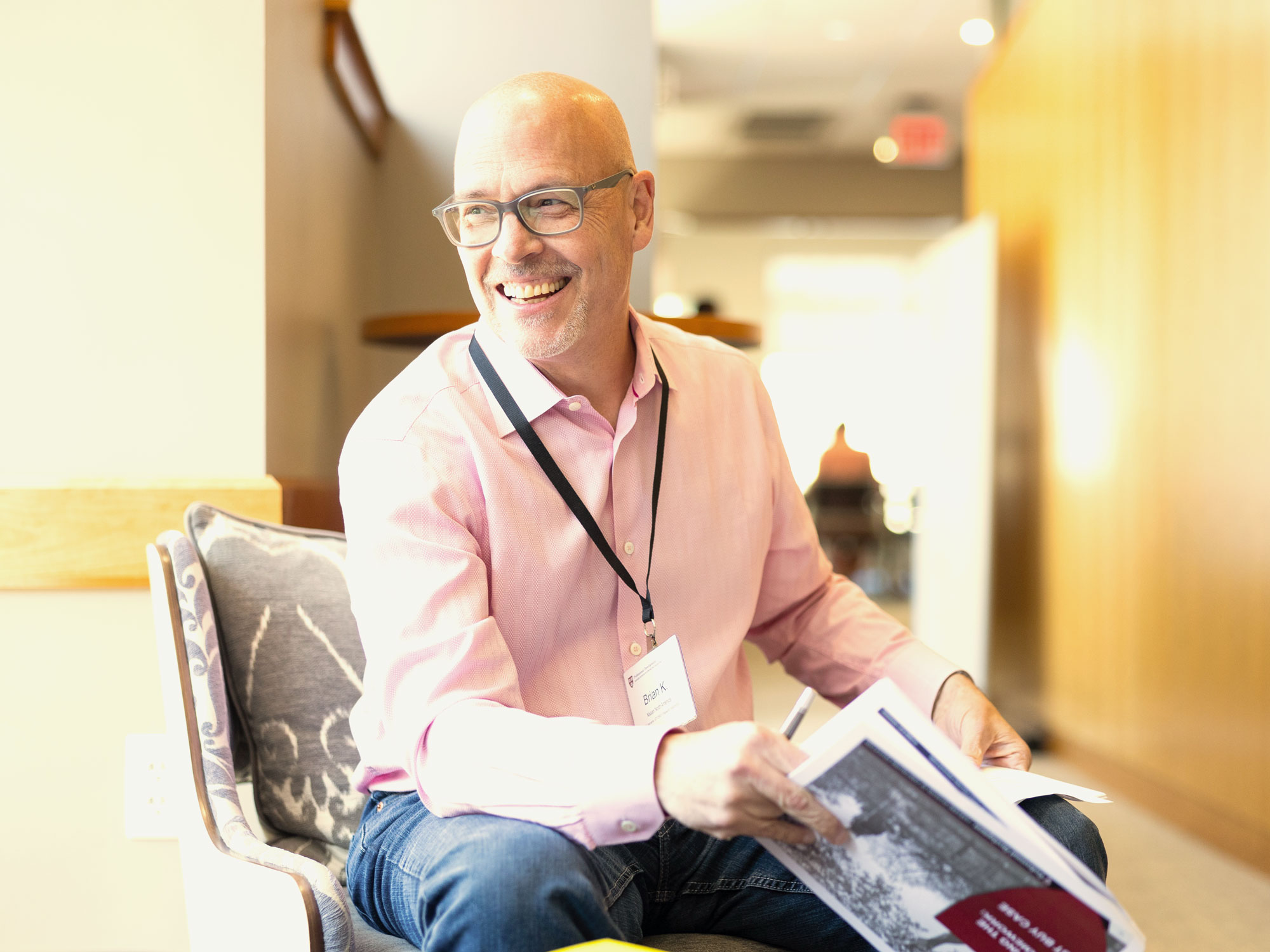 A sitting man in a pink dress shirts smiles at someone out of frame while holding documents