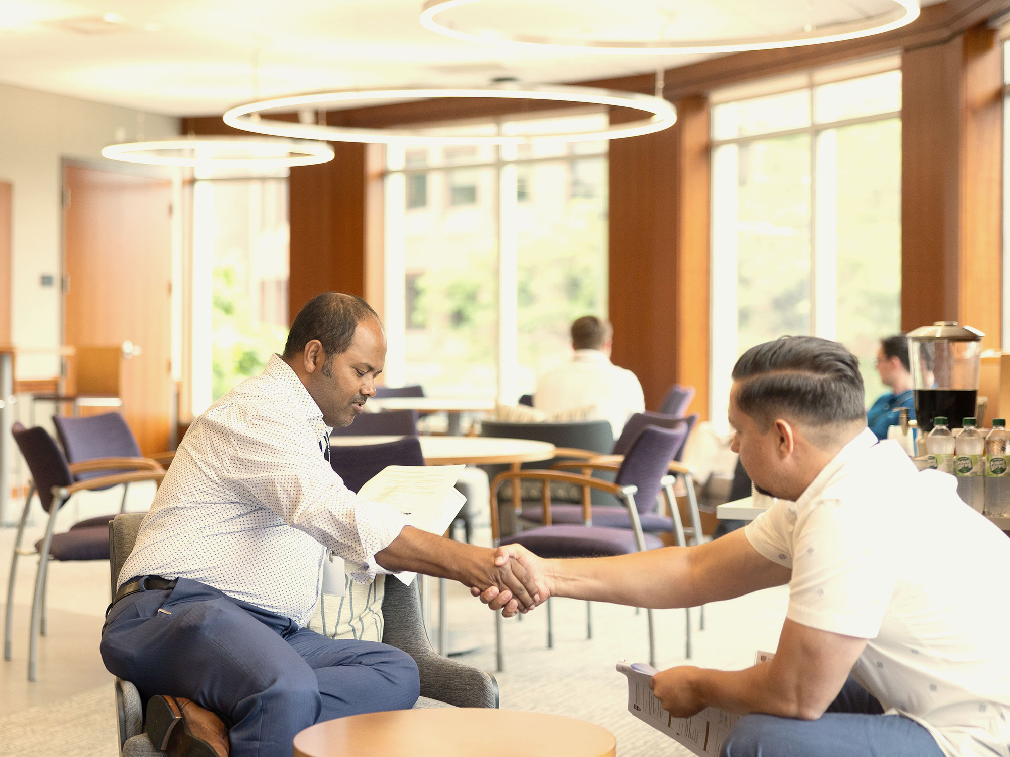 Two men in business attire shaking hands while sitting near large windows in a bright meeting space.